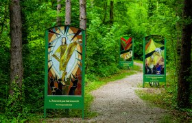 Weg durch einen Wald, links und rechts neben dem Weg stehen gro&szlig;e Tafeln aus Glas, die die Stationen des Kreuzweges darstellen.