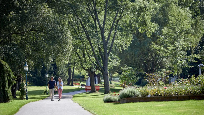 Ein Paar spaziert auf einem Weg im Kurpark Bad Schönau, umgeben von grünen Bäumen und gepflegten Blumenbeeten.