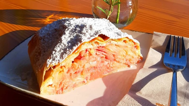 Ein Stück Apfelstrudel mit Puderzucker auf einem Teller, daneben eine Gabel und eine Vase mit Rosmarin.