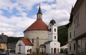 Pfarrkirche Scheiblingkirchen mit rundem Turm und rotem Dach, umgeben von Gebäuden und bewölktem Himmel.