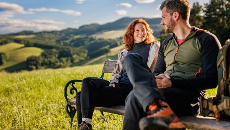 Eine Frau und ein Mann in Wanderkleidung sitzen auf einer Holzbank. Im Hintergrund Wiese, grüne Hügel und Wald.