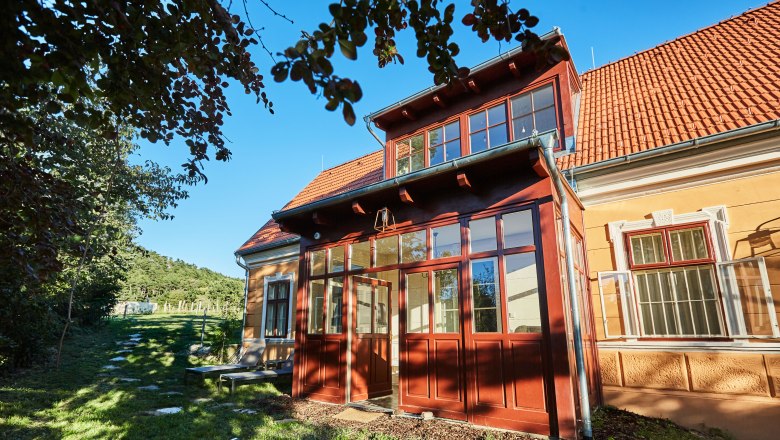 Eine gelbe Villa mit roten Dachziegeln und einem verglasten Eingang, umgeben von Bäumen und einem grünen Garten.