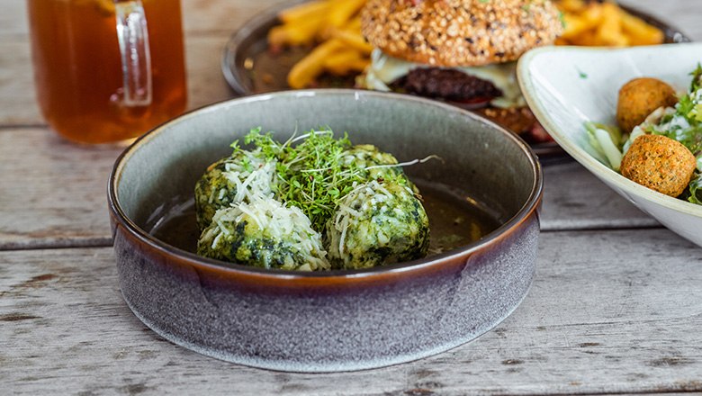 Ein Teller mit Spinatknödeln, Parmesan und Kresse. Im Hintergrund ein Getränk, ein Burger und ein Salat.