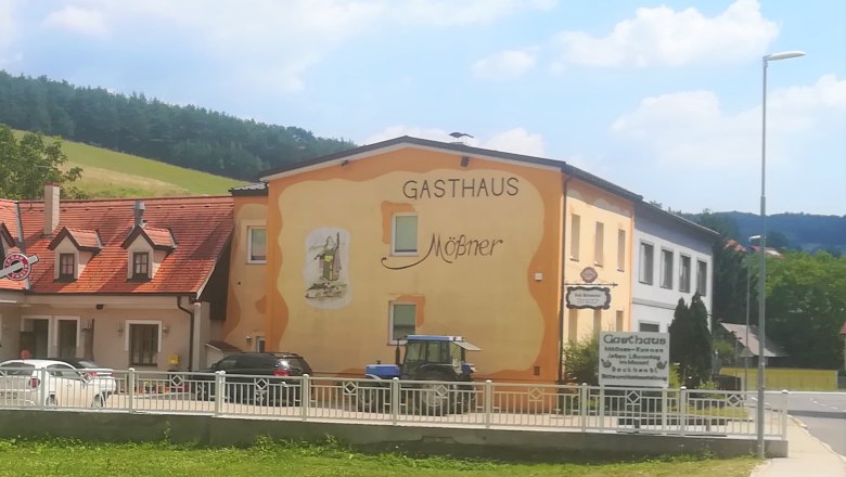 Exterior view of the Mößner inn, painted yellow-orange, in a rural setting with a blue tractor and cars in front of the building.