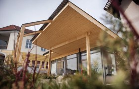 Modernes Gebäude mit Holzüberdachung und Glasfront, umgeben von Pflanzen.