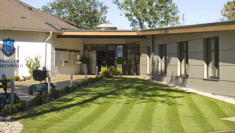 Eingang des Clubhauses des Golfclubs F&ouml;hrenwald mit gepflegtem Rasen und modernen Geb&auml;uden.