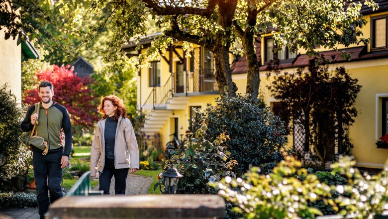 A man and a woman in hiking gear walk through the green, well-tended garden of the Pension Hendling