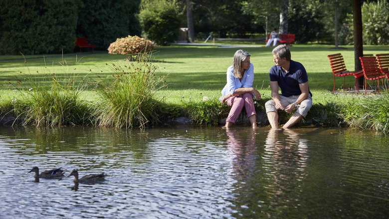 Ein Paar sitzt am Ufer eines Teichs im Kurpark Bad Sch&ouml;nau, die F&uuml;&szlig;e im Wasser. Zwei Enten schwimmen im Vordergrund.