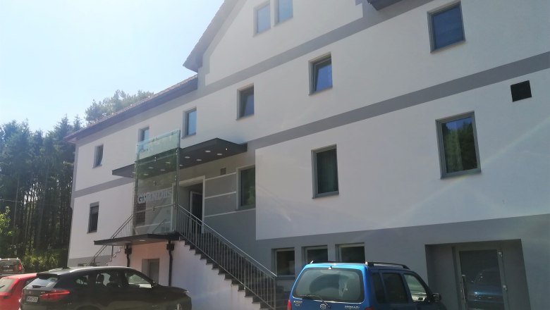 Außenansicht eines modernen Gasthauses mit mehreren Fenstern und parkenden Autos davor.