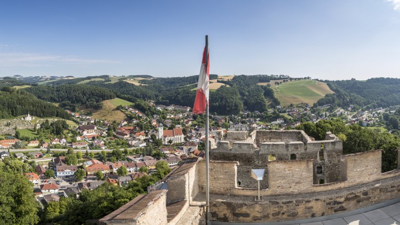 Panoramablick von einer Burgruine auf eine Stadt mit Hügeln im Hintergrund und einer österreichischen Flagge im Vordergrund.