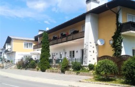 Ein zweistöckiges Gebäude mit Balkon und Terrasse, umgeben von Pflanzen und Bäumen, bei sonnigem Wetter.