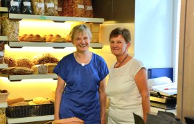 Zwei Frauen in einer Bäckerei vor Regalen mit Brot und Gebäck.