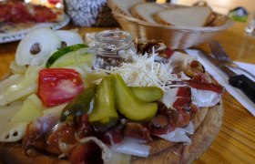 Ein rustikales Brett mit K&auml;se, Schinken, Tomaten, Gurken und Zwiebeln, daneben ein Brotkorb.