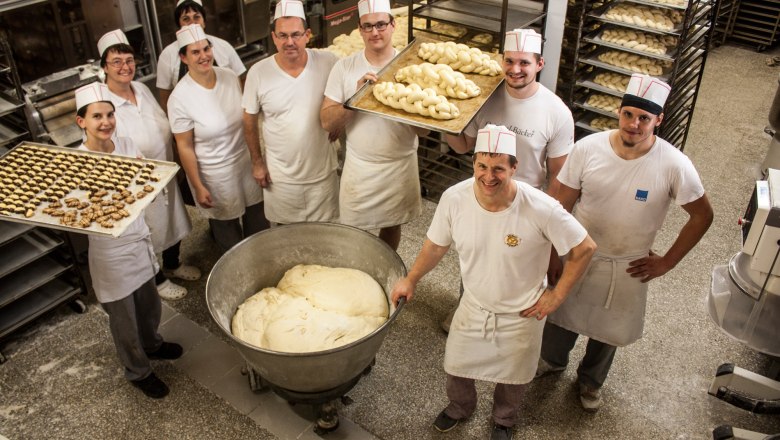 Mitarbeiter einer Bäckerei in weißer Arbeitskleidung mit Blechen voll Teigwaren in einer Backstube.