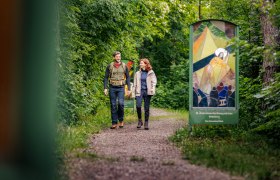 Zwei Personen wandern auf einem Waldweg mit gläsernen Kunstwerken am Rand.
