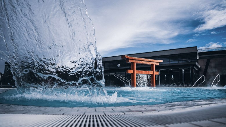Ein Torii-Tor in einem Pool mit sprudelndem Wasser und einem modernen Gebäude im Hintergrund.