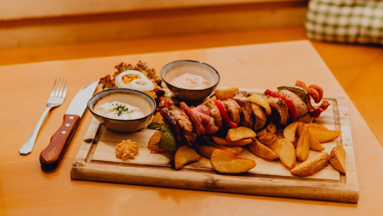 Ein Holzbrett mit einem Fleischspieß, Kartoffelecken, zwei Saucenschalen und Salat auf einem Tisch.