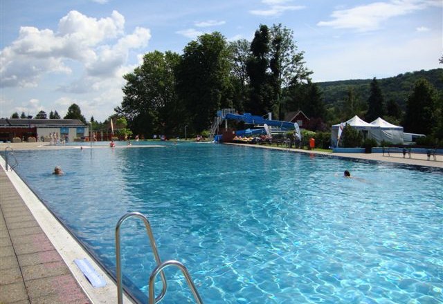 A large outdoor pool with clear water, surrounded by trees and hills under a blue sky.