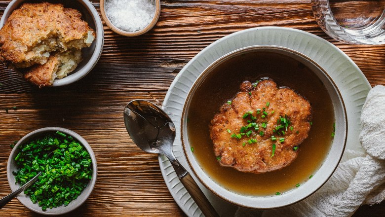 Kaspressknödelsuppe in einer Schüssel auf einem Holztisch, mit Schnittlauch und Salz in kleinen Schalen daneben.