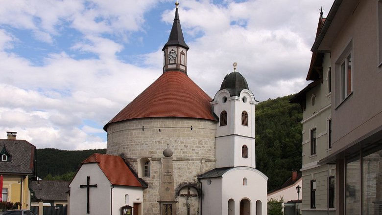Pfarrkirche Scheiblingkirchen mit rundem Turm und rotem Dach, umgeben von Gebäuden und bewölktem Himmel.
