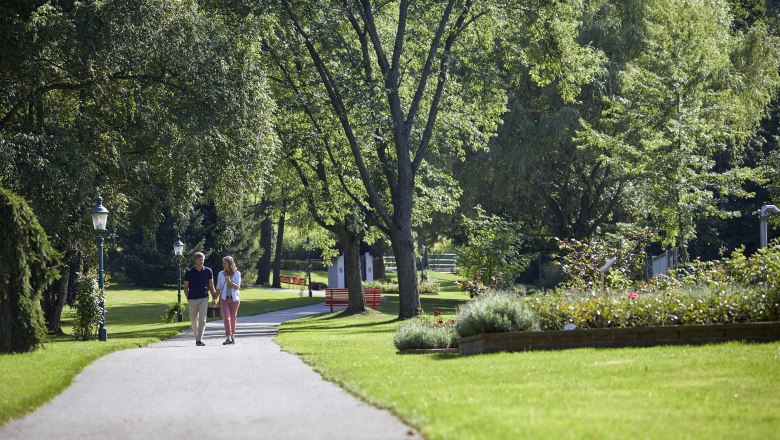 A couple walks along a path in the spa gardens of Bad Schönau, surrounded by green trees and well-tended flowerbeds.