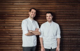 Two men in white shirts stand in front of a wooden wall and smile.
