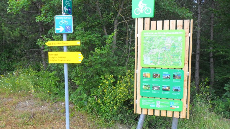 Wegweiser und Informationstafel f&uuml;r Radfahrer in Bad Erlach, umgeben von B&auml;umen.