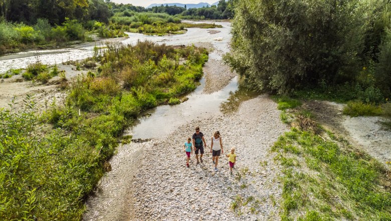 Eine Familie spaziert durch eine grüne Flusslandschaft mit Kieselsteinen und Bäumen. Luftaufnahme