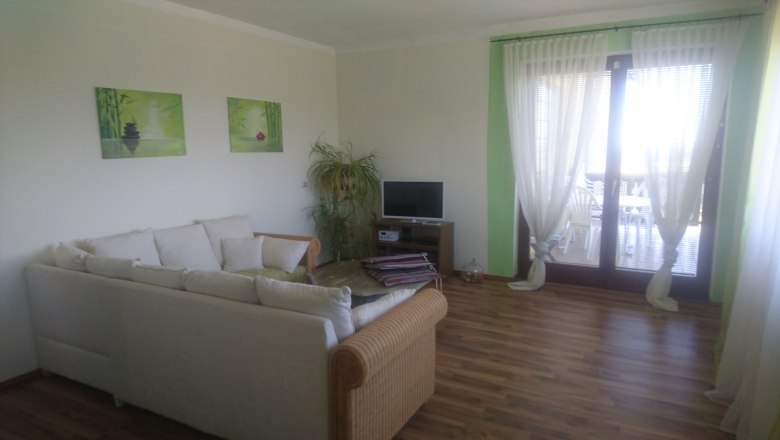 A living room with a white sofa, a small TV, plants and large windows with curtains.
