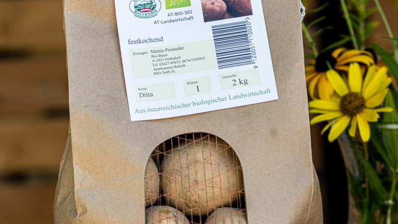 Ein Papiersack mit Bio Erdäpfeln, daneben gelbe Blumen.