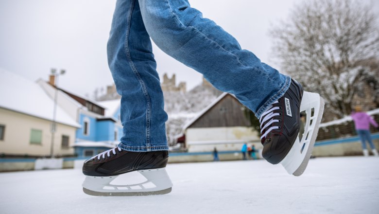 Nahaufnahme von Schlittschuhen auf einer Eisfläche mit Gebäuden und Burgruine im Hintergrund.
