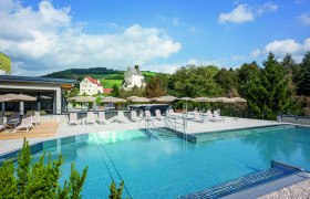 Außenpool mit Liegestühlen und Sonnenschirmen, im Hintergrund eine Kirche und grüne Hügel.