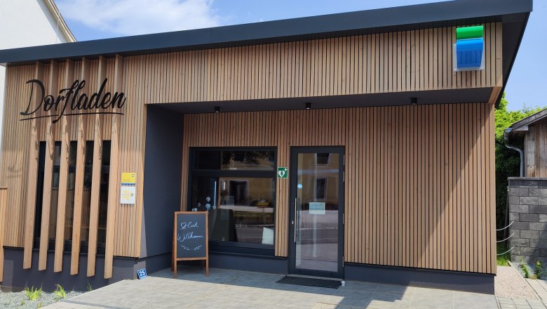 Exterior view of a modern village store with wooden paneling and a welcome sign in front of the door.