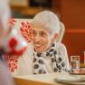 Elderly woman sitting at a table smiling, in the foreground a cup with a floral pattern and a glass of water.
