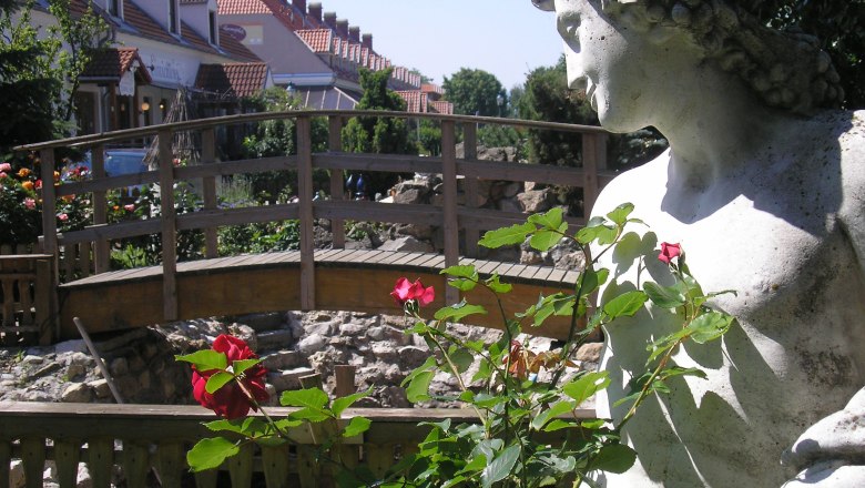 Steinstatue neben einer Holzbr&uuml;cke und roten Rosen im Vordergrund.