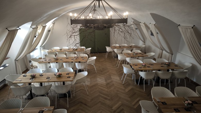 Eleganter Saal mit Holztischen, Parkettboden, wei&szlig;en St&uuml;hlen und eisnerner Kronleuchter.