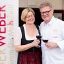 Ein Mann und eine Frau stoßen mit Weingläsern an, neben einem Schild mit der Aufschrift 'Der Weber'.