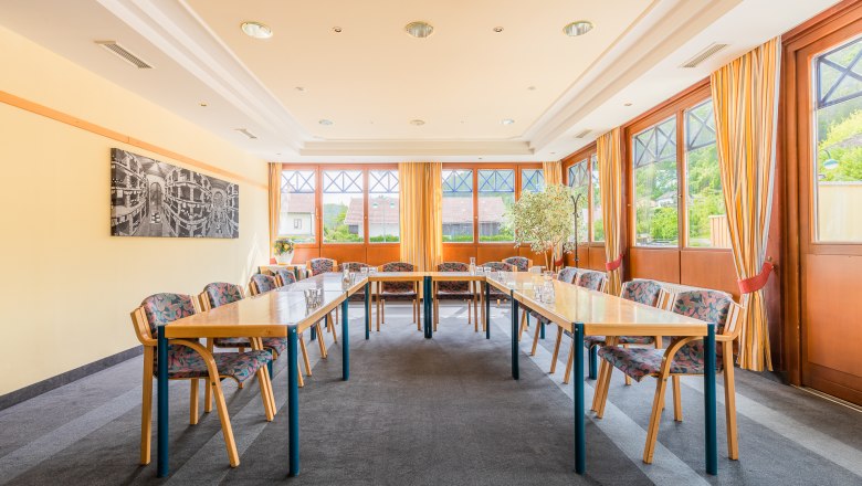 Ein heller Seminarraum mit Tischen in U-Form, Stühlen und großen Fenstern mit Vorhängen.