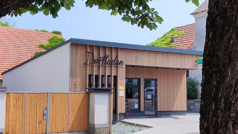A modern village store with a wooden façade and signs, surrounded by trees.