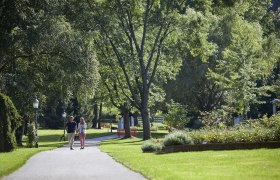 Ein Paar spaziert auf einem Weg im Kurpark Bad Schönau, umgeben von grünen Bäumen und gepflegten Blumenbeeten.