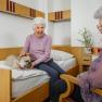 Two older women in a room, one petting a dog on the bed.