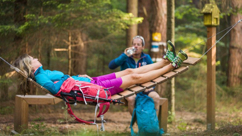Zwei Personen entspannen im Wald, eine liegt in einer Hängematte, die andere trinkt aus einer Flasche.