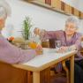 Two elderly women are sitting at a table in a cozy room. One woman is pouring juice from a carafe into a glass.