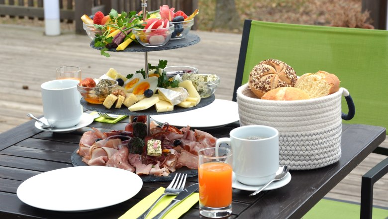 Frühstückstisch im Freien mit Aufschnitt, Käse, Gemüse. Daneben Teller, Besteck, Getränke und Brotkorb.