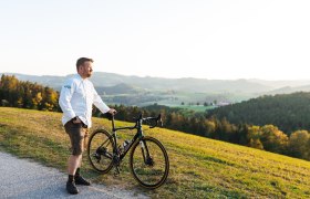 Ein Mann in wei&szlig;em Hemd steht mit einem Fahrrad auf einem H&uuml;gel mit Blick auf eine gr&uuml;ne Landschaft.