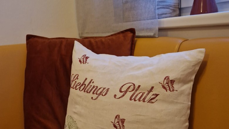 Ein gemütliches Sofa mit einem dekorativen Kissen, auf dem 'Lieblings Platz' steht, neben einem Fenster mit Pflanzen und einer Vase.