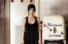 Eine Frau in einem schwarzen Kleid steht neben einer alten B&auml;ckereimaschine mit der Aufschrift 'Fortuna'.