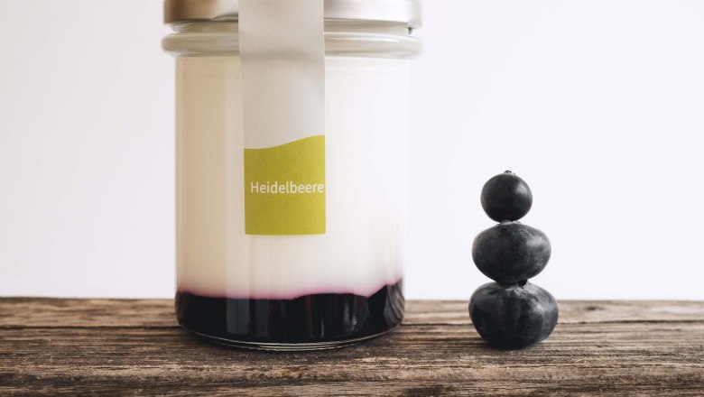 Ein Glas gef&uuml;llt mit Joghurt und Fruchtspiegel auf einem Holztisch, daneben 3 gestapelte Heidelbeeren.
