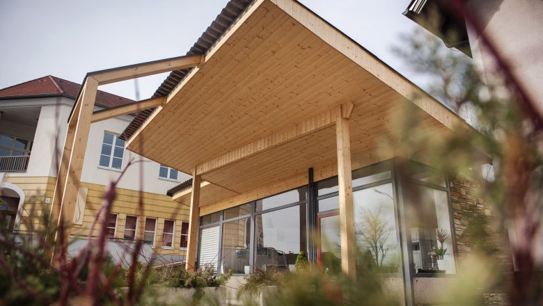 Modernes Gebäude mit Holzüberdachung und Glasfront, umgeben von Pflanzen.