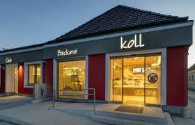 Au&szlig;enansicht am Abend der B&auml;ckerei mit beleuchtetem Schild 'B&auml;ckerei Koll' und 'Cafe'.